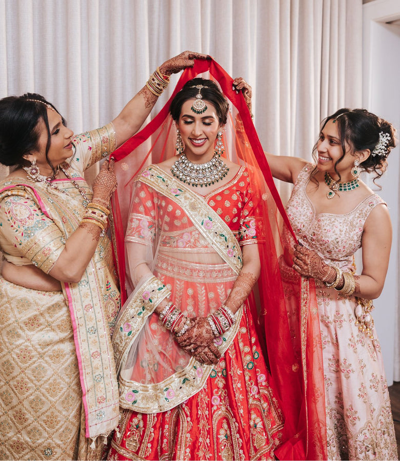Indian bride in a red bridal lehenga wearing a layered necklace, earrings, and maang tikka, flanked by two women adjusting her veil