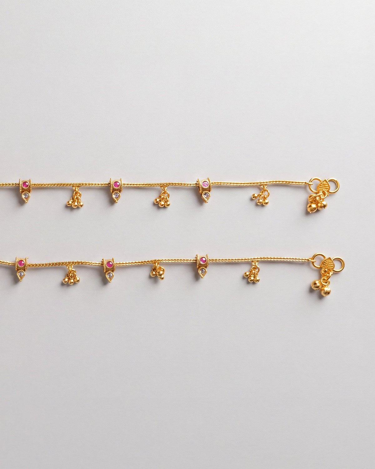 Flat lay detail of matching Mira Dewdrop Anklets, gold-tone chain with tiny bells, clear pear stones, and pink accents